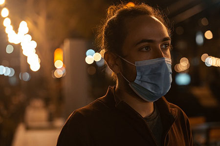 Foto de uma pessoa com máscara de proteção contra Covid-19, andando na rua a noite, com luzes ao fundo